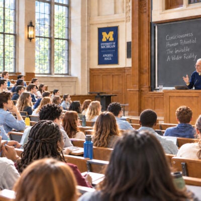 A man with a gray ponytail and glasses and a podium before an audience of students